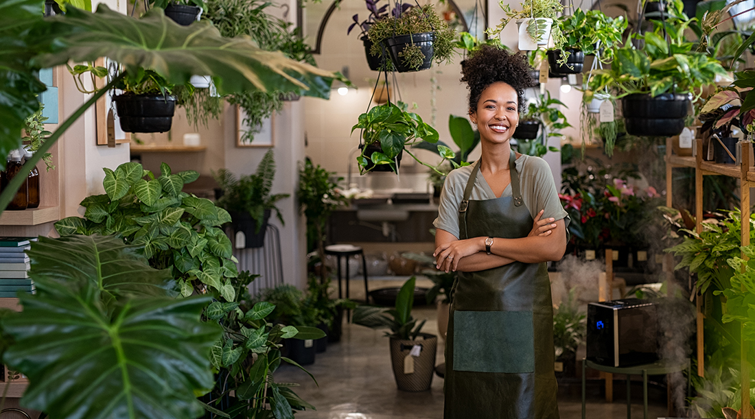 Portrait of african american woman with crossed arms wearing apron standing in botanical store. Smiling young woman in botany store standing between plants looking at camera. Happy small business owner working at flower shop standing surrounded by plants.